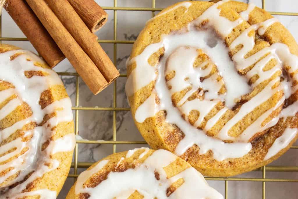 Close-up of pumpkin cinnamon roll cookies drizzled with white maple icing, resting on a gold cooling rack. A few cinnamon sticks are placed to the side, emphasizing the warm, spiced flavor of the cookies