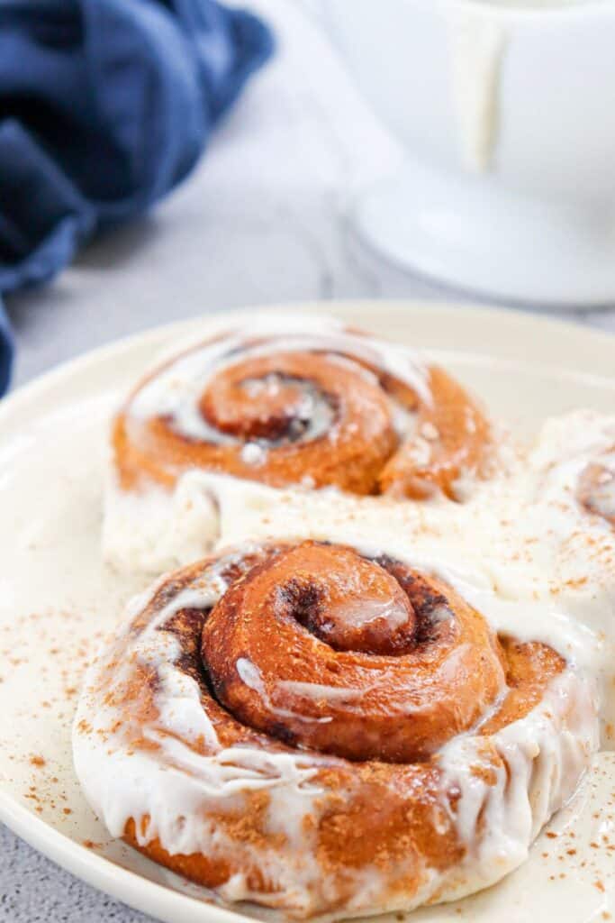 Plate of two iced pumpkin cinnamon rolls with glass jug of milk, and tea towel in background.