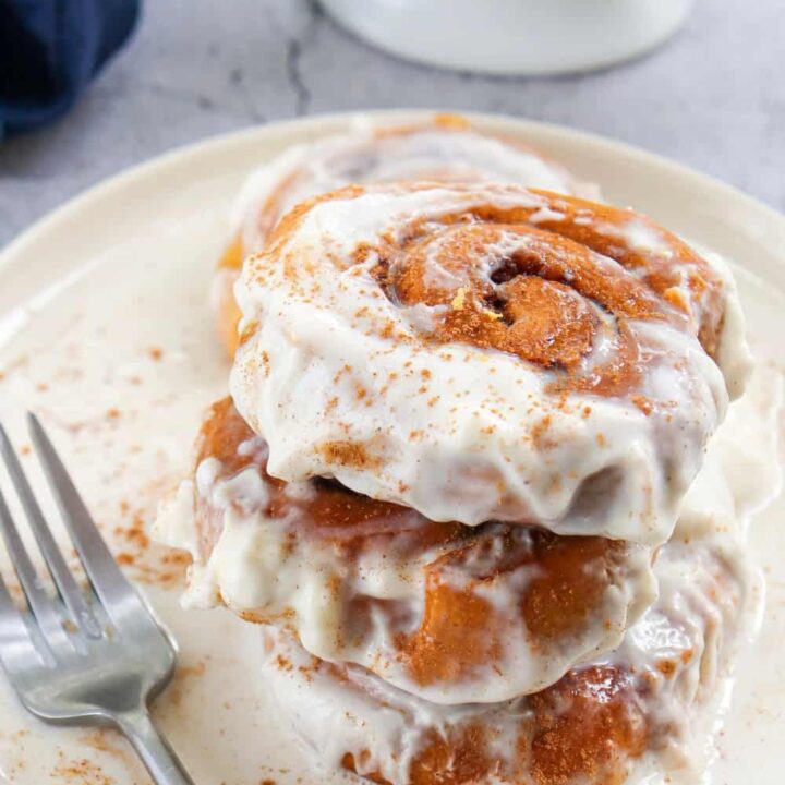 Pile of three iced pumpkin cinnamon rolls on a plate set on marbled table with a teat towl in background.