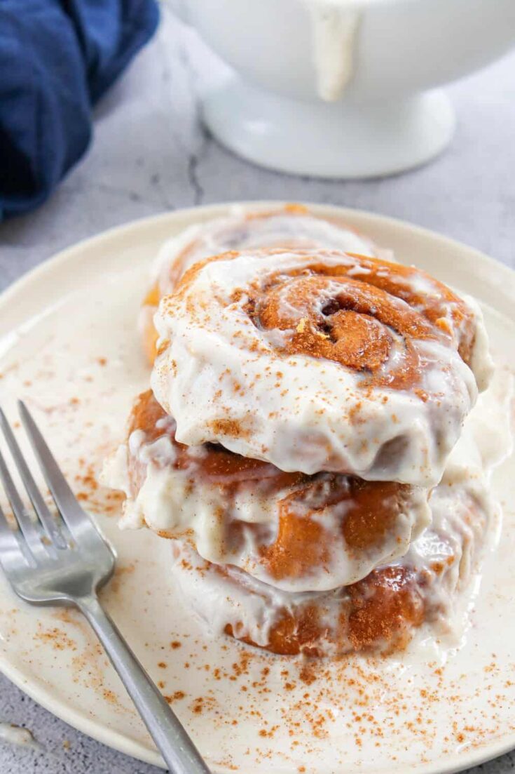 Pile of three iced pumpkin cinnamon rolls on a plate set on marbled table with a teat towl in background.