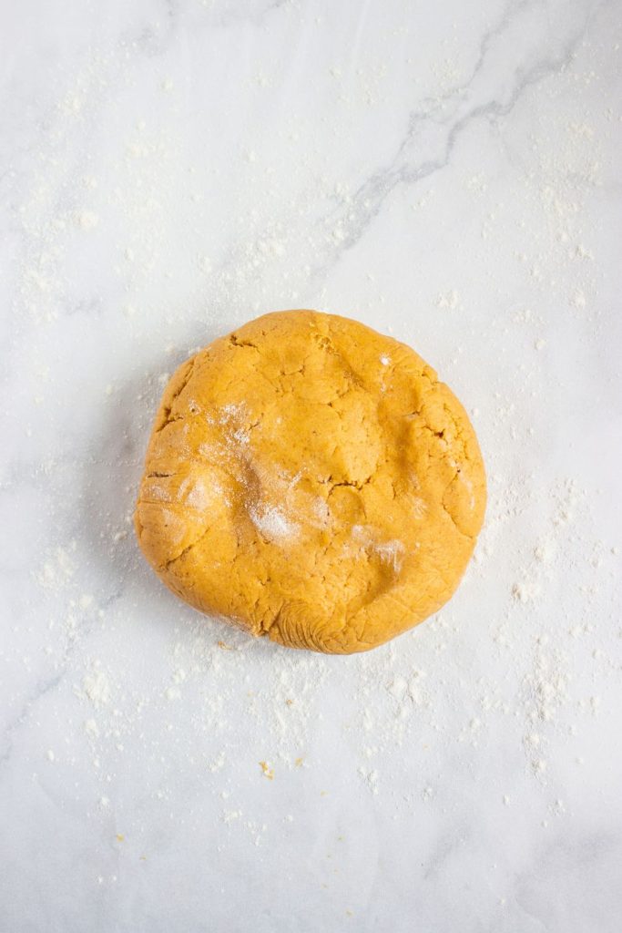 A ball of pumpkin cookie dough resting on a lightly floured marble surface, slightly dusted with flour and ready to be rolled out. The dough has a warm orange hue and smooth, pliable texture.