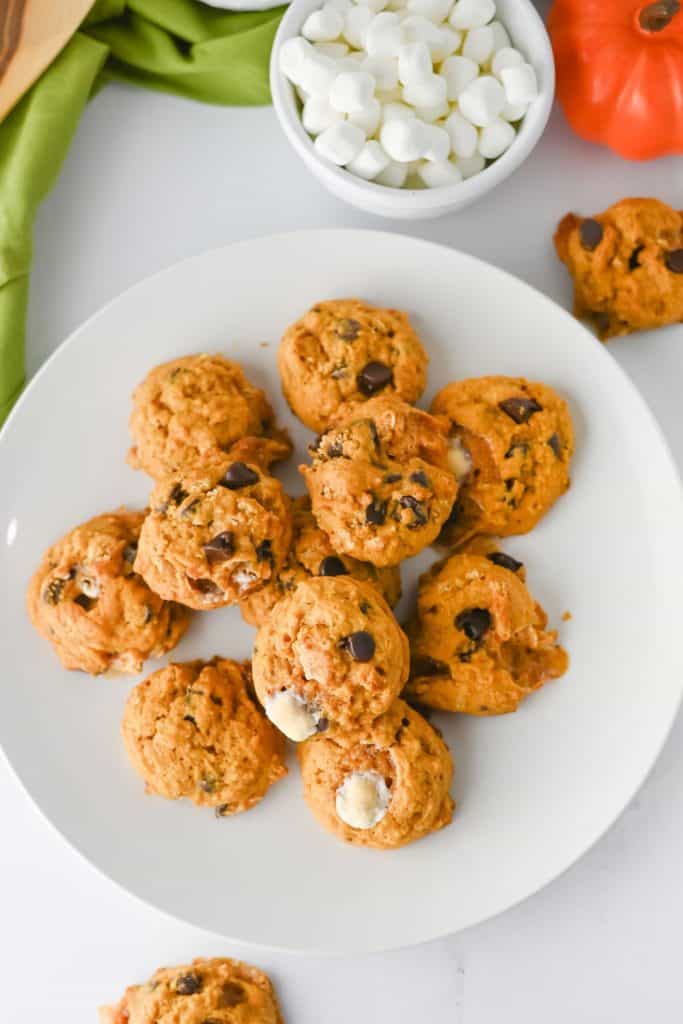 Overhead view of freshly baked cookies on a white plate, filled with chocolate chips and melted marshmallows. A bowl of mini marshmallows and an orange decorative pumpkin sit nearby, adding a cozy, fall-inspired touch.