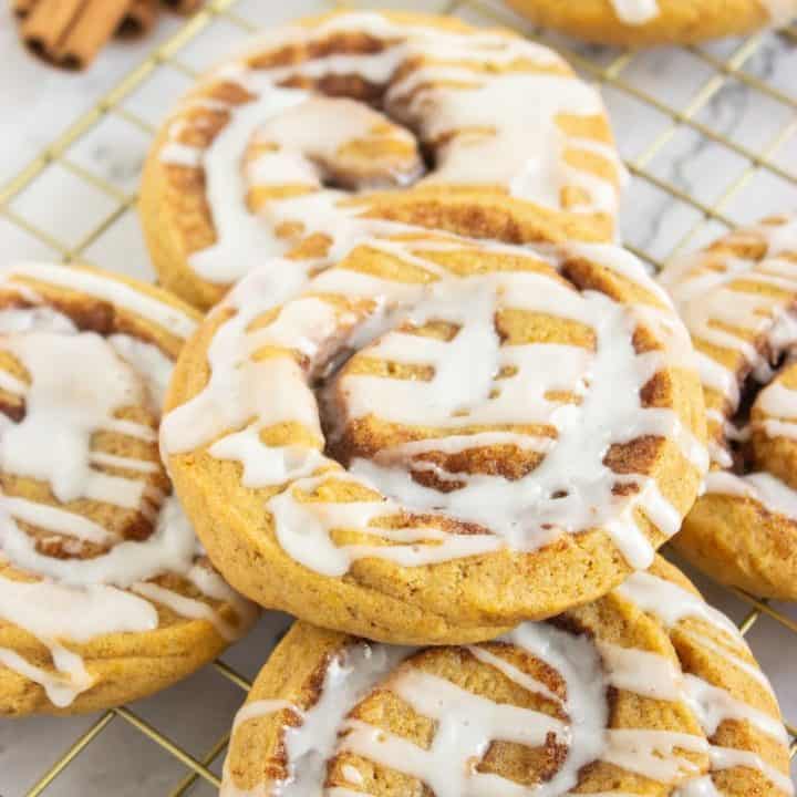 A stack of pumpkin cinnamon roll cookies drizzled with white icing, cooling on a wire rack. The cookies are golden brown with visible cinnamon swirls, and a bundle of cinnamon sticks is tied with burlap ribbon in the background.