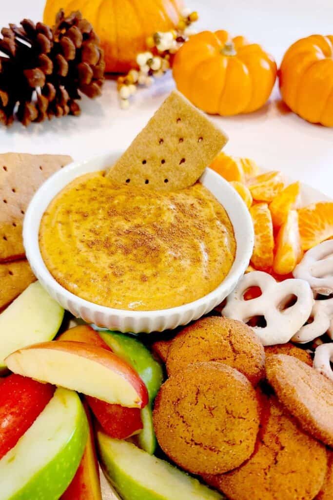 Small bowl of pumpkin cream cheese dip surrounded by pretzles, apple slices and gingersnap cookies. 