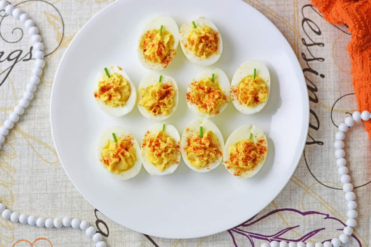 Pumpkin deviled eggs arranged on a white plate with yellow filling dusted with paprika. Small green chive stems give each egg a pumpkin like appearance.