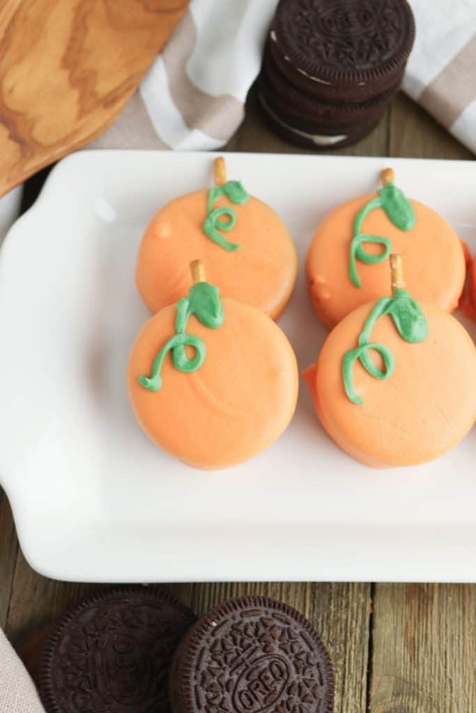 Oreo cookies dipped in orange candy coating to resemble pumpkins, topped with green icing vines and a small pretzel stick stem. Displayed on a white tray with extra plain Oreos nearby, creating a festive fall or Halloween treat.