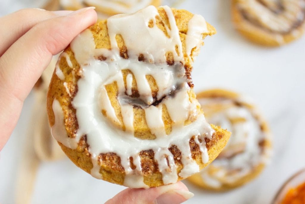 A hand holding a soft pumpkin cinnamon roll cookie with a bite taken out, showing its gooey cinnamon swirl and white icing drizzle. More cookies are slightly blurred in the background on a light surface for perfect fall baking ideas. 