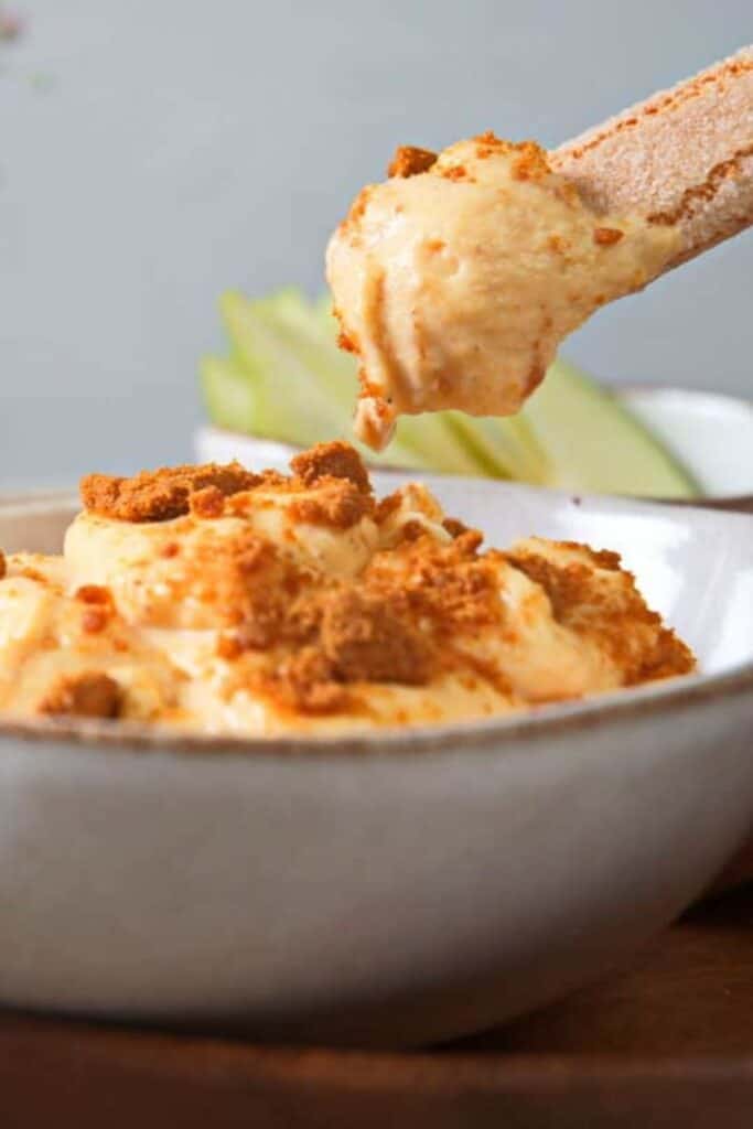 Pumpkin fluff topped with crumbled cookies in a white bowl, with a cinnamon sugar cookie scooping up the creamy dip. This easy treat is served alongside green apple slices and works well as fall potluck desserts.