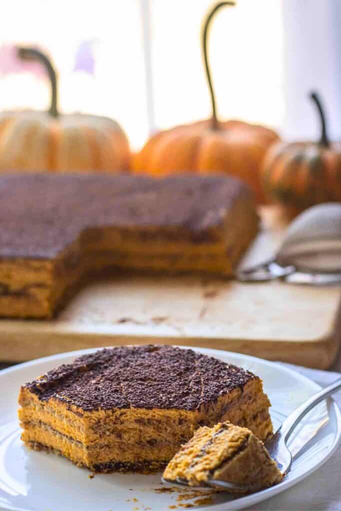 Slice of pumpkin ice box cake with creamy spiced pumpkin layers and thin graham cracker layers topped with a dusting of cocoa. A spoonful beside the slice shows the soft mousse like texture of the no bake fall pumpkin dessert.
