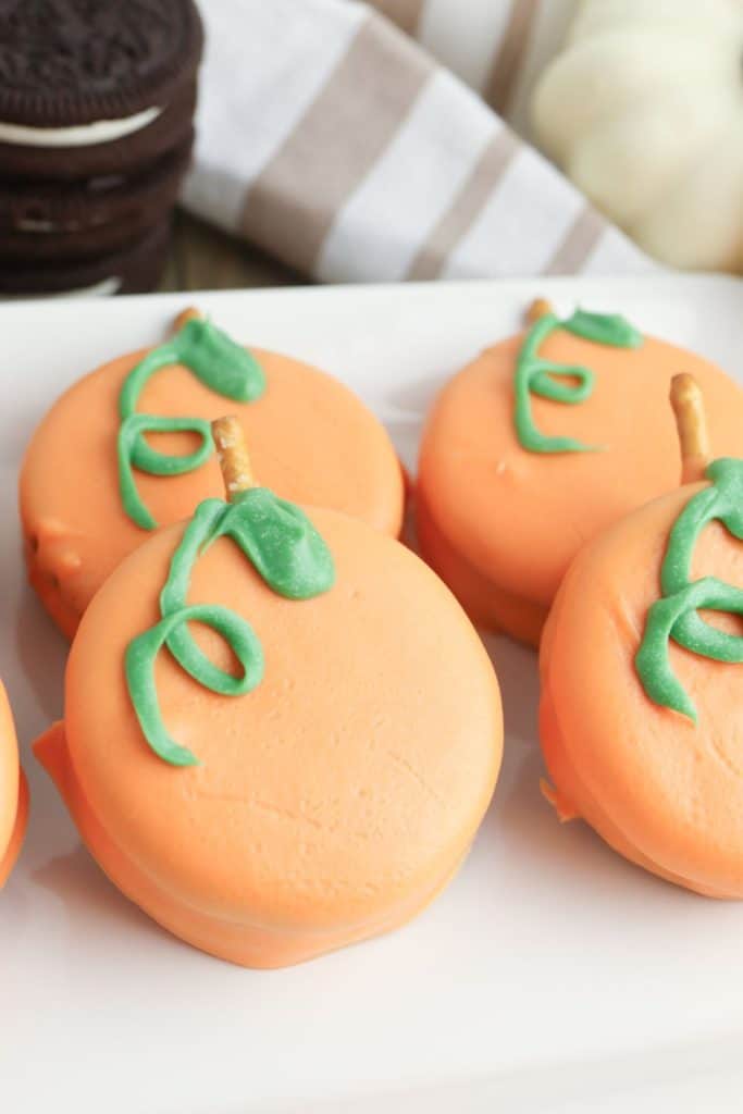 Close-up of four orange candy-coated cookies decorated to look like pumpkins, with green icing vines and pretzel stick stems. The treats sit on a white plate with stacked chocolate sandwich cookies and white pumpkins blurred in the background.