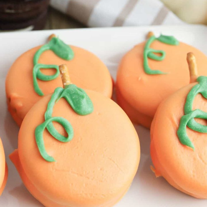 Close-up of four orange candy-coated cookies decorated to look like pumpkins, with green icing vines and pretzel stick stems. The treats sit on a white plate with stacked chocolate sandwich cookies and white pumpkins blurred in the background.