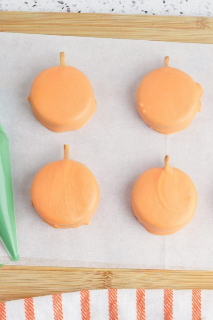 Four orange candy-coated cookies with pretzel stick stems are arranged on parchment paper atop a wooden board. A green piping bag sits on the side, and an orange and white striped towel is partially visible at the bottom of the frame.