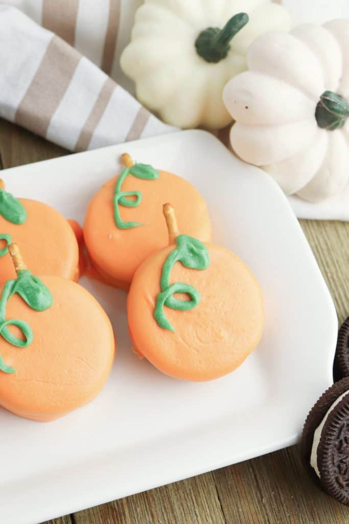Plate of Oreo Cookies, decorated to look like pumpkins with orange candy coating, green icing vines, and pretzel stick stems. White decorative pumpkins and striped linens complete the festive fall presentation.