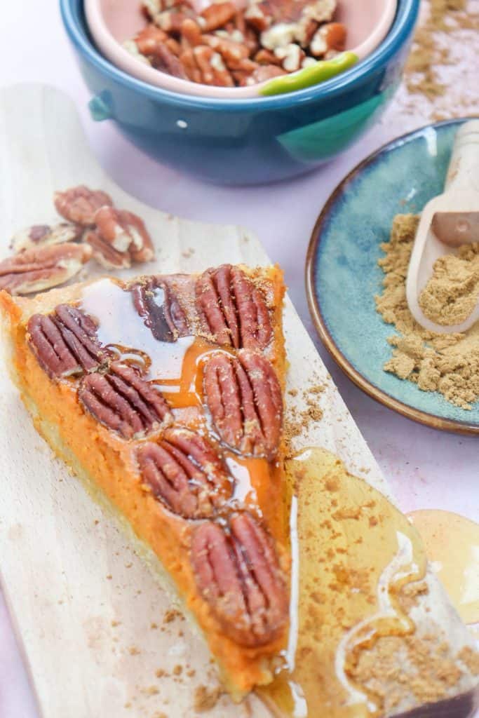 A slice of pumpkin pecan pie topped with whole glazed pecans and drizzled with syrup, resting on a wooden board. Surrounding it are raw pecans, brown sugar on a ceramic plate, and a colorful bowl filled with more nuts.