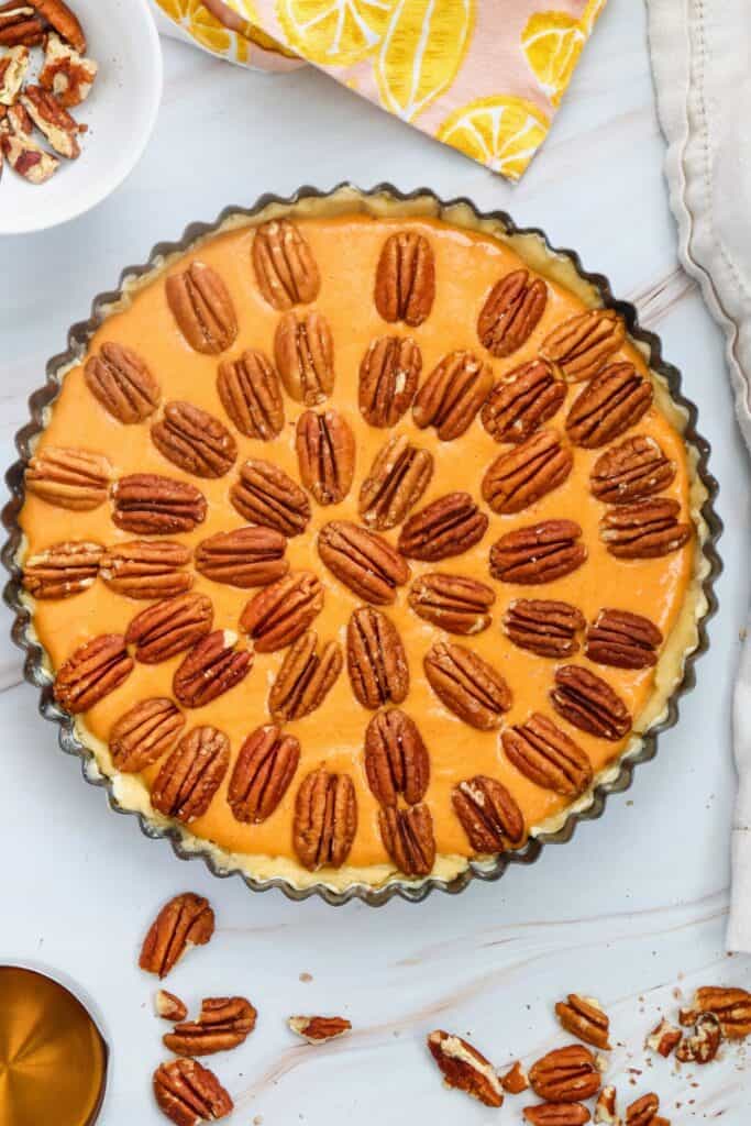 Overhead view of a Pumpkin Pecan Cheesecake with creamy orange pumpkin filling topped with whole pecans in a fluted crust, surrounded by scattered pecans.