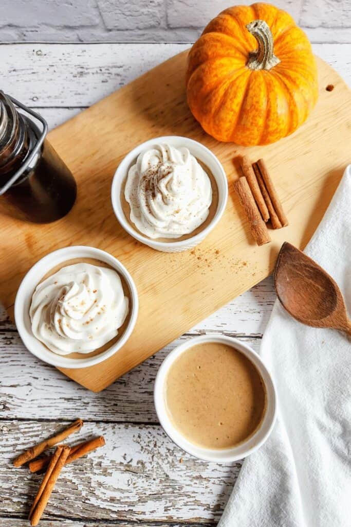 Three bowls of pumpkin pie pudding cups, with two of them topped with whipped cream, set on a wood cutting board beside cinnamon sticks and small pumpkin.