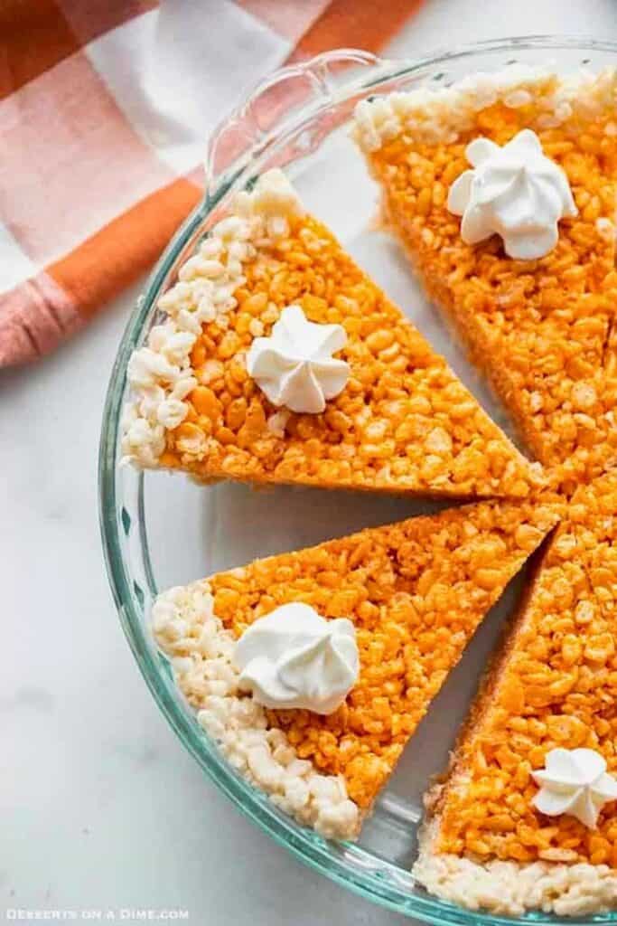 Fall Rice Krispie treats shaped like slices of pumpkin pie with orange cereal centers and white cereal crusts. Each wedge is topped with a dollop of whipped cream and arranged in a glass pie dish.