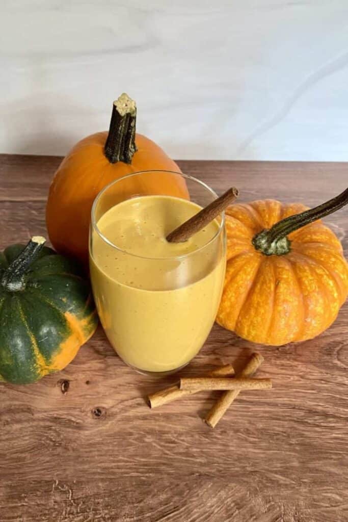 A clear glass of pumpkin pie smoothie beside 3 small fall gourds. 