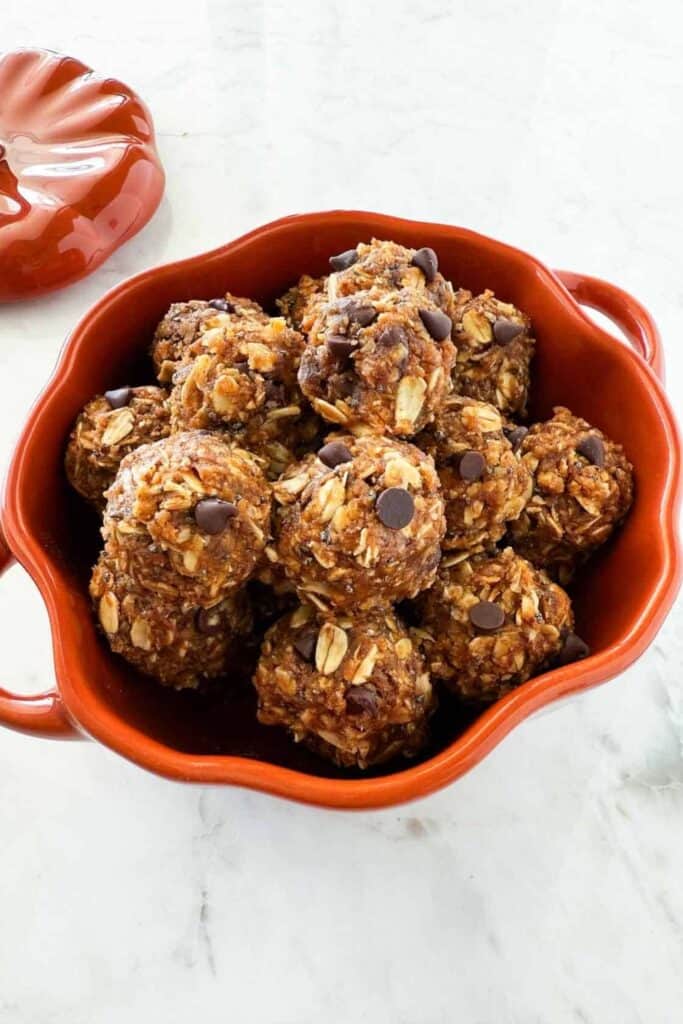 umpkin protein balls made with rolled oats and chocolate chips piled in an orange serving bowl. The bite sized no bake treats have a textured oat coating and warm pumpkin spice color.
