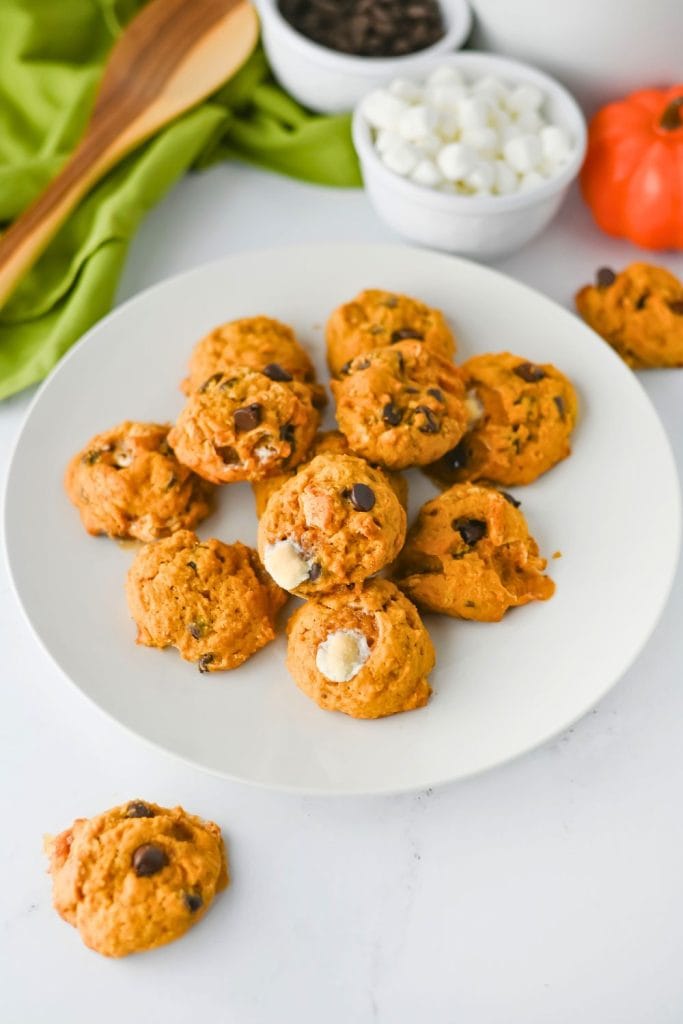 Plate of soft and fluffy pumpkin s’mores cookies made with chocolate chips and gooey melted marshmallows. Surrounding the dish are mini marshmallows, chocolate chips, and a decorative pumpkin, creating a cozy fall baking scene.