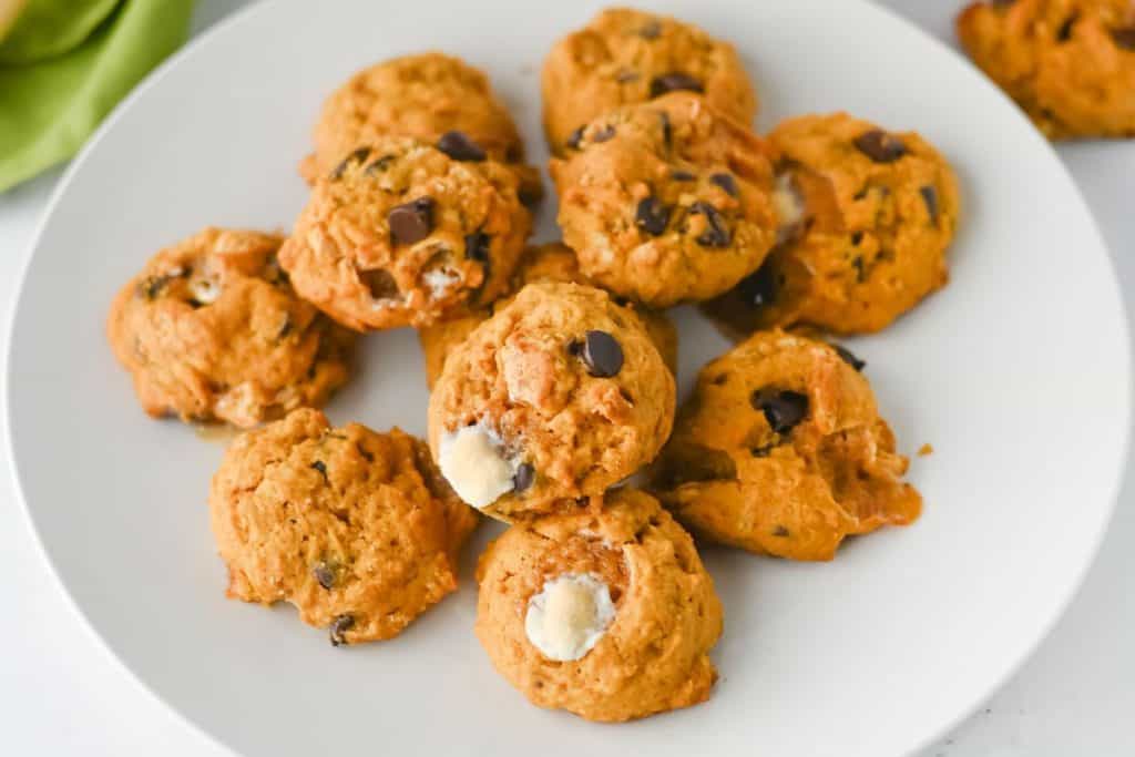 Close-up of a white plate filled with pumpkin cookies studded with chocolate chips and gooey marshmallows. The cookies have a soft, golden-orange texture, showcasing cozy, homemade fall treats.
