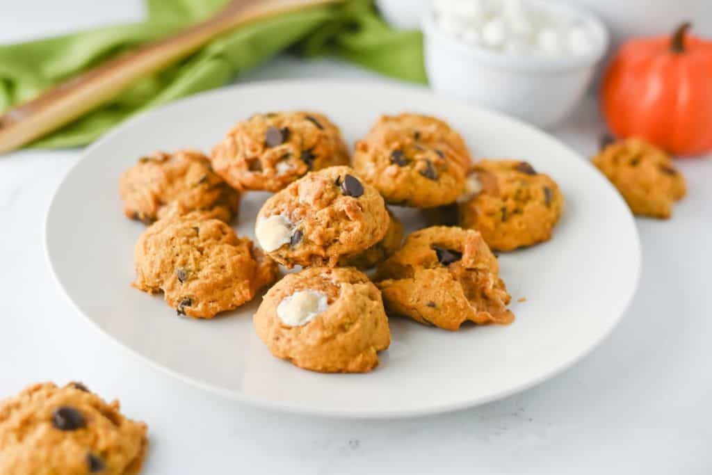 A plate of pumpkin s’mores cookies with visible chocolate chips and melted marshmallows, styled on a white surface with a mini pumpkin and bowl of marshmallows in the background. These soft, fall-themed cookies blend pumpkin flavor with classic s’mores ingredients.
