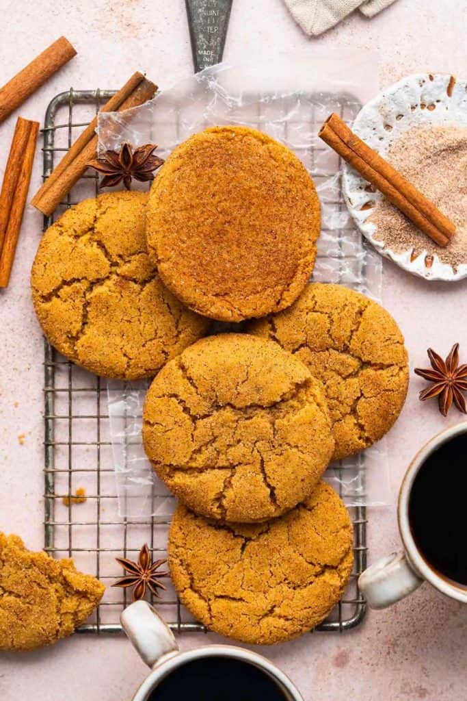 Golden-brown Pumpkin Snickerdoodles stacked on a cooling rack, their cracked tops dusted with cinnamon sugar. Surrounded by cinnamon sticks, star anise, and cups of coffee, the scene evokes cozy fall baking vibes.