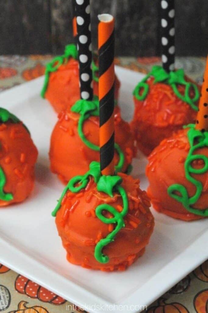 Pumpkin spice cake pops coated in bright orange candy and decorated with green icing vines and leaves. Black and orange striped paper straws serve as stems for the festive pumpkin shaped treats.