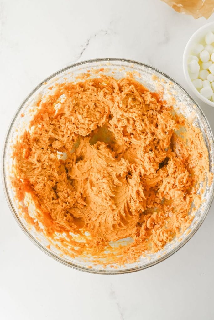 Overhead view of a glass bowl filled with thick, orange cookie dough, freshly mixed and ready for baking. A bowl of mini marshmallows and a graham cracker wrapper are partially visible nearby, suggesting a sweet and seasonal recipe.