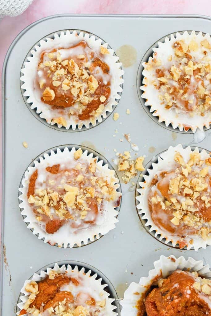 Pumpkin walnut muffins baked in white paper liners, drizzled with icing and topped with chopped walnuts. Shown in a muffin tin with a few crumbs and drips, the warm, spiced treats highlight cozy fall movie night snacks.