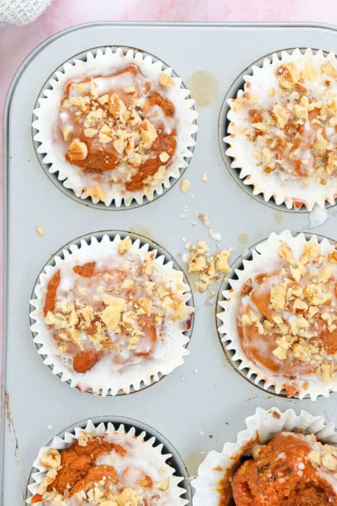 Muffin tin of six iced pumpkin muffins topped with walnut pieces. 