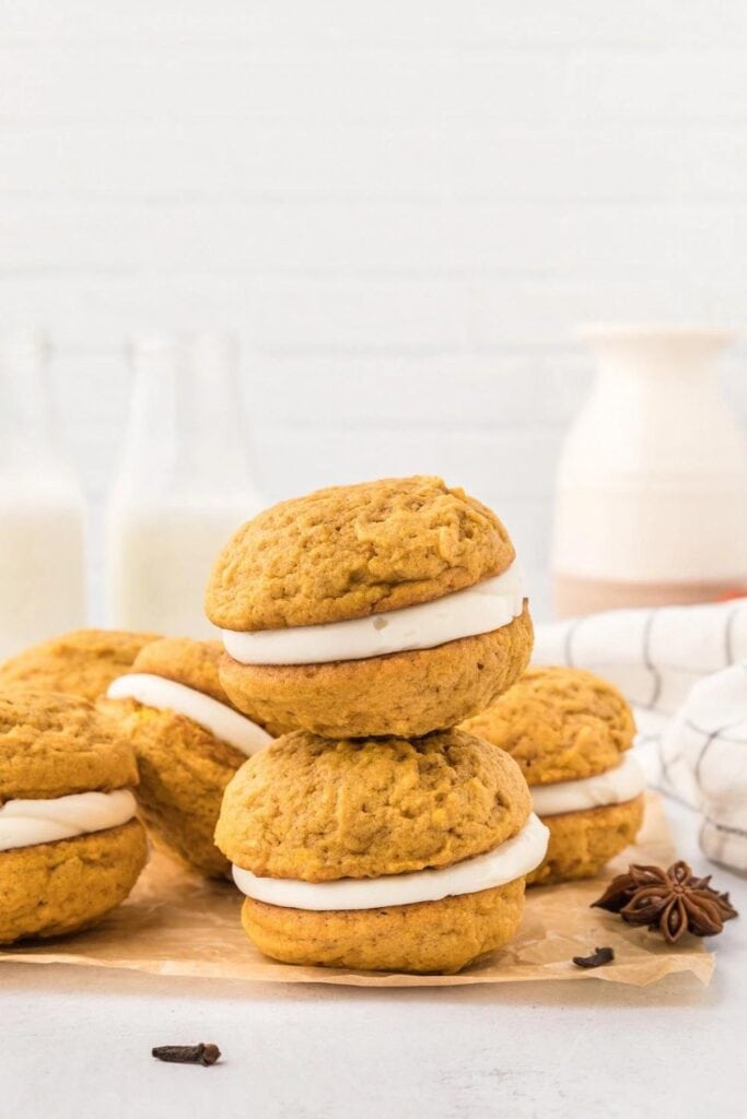 6 Pumpkin whoopie pies with cream cheese icing are scattered on parchment paper, in front of glass jars of milk.