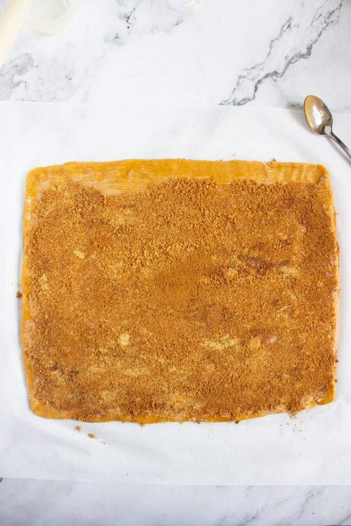 Rolled-out pumpkin cookie dough evenly coated with a layer of brown sugar and cinnamon mixture, spread to the edges. A spoon and bits of extra filling rest nearby on a marble surface, showing the dough prepped and ready to roll into cinnamon roll style cookies.