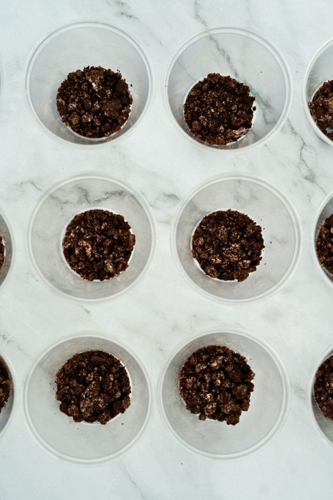 Overhead view of clear plastic cups partially filled with crushed chocolate cookies, arranged in a grid on a marble surface as the base layer for no bake Halloween treats.