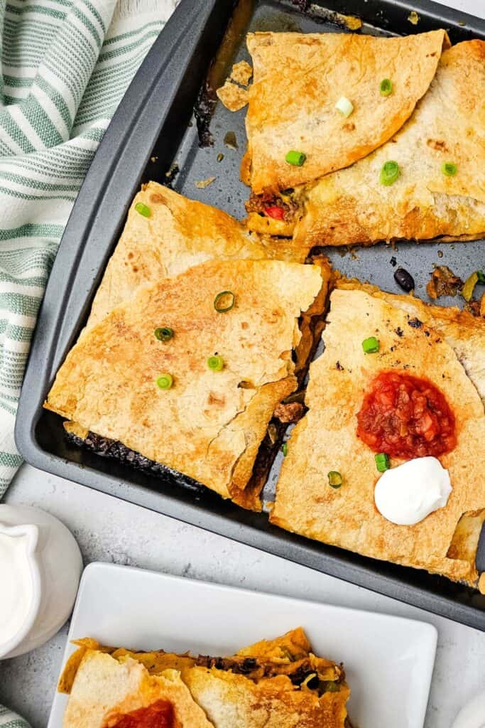 Baked sheet pan quesadillas cut into crispy squares, with some topped with sour cream, salsa, and chopped green onions. Served alongside a dipping dish of sour cream, this easy tortilla-based meal is perfect for quick dinner ideas.