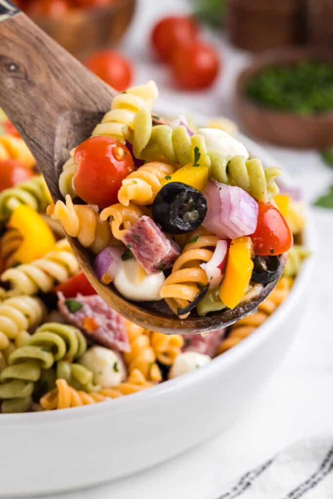 Close up of a wooden spoon full of spiral pasta salad with red onion, cherry tomatoes, olives, peppers and cheese. 