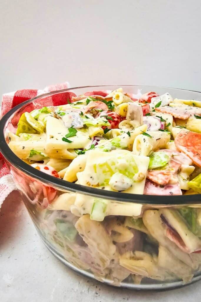 Side view of a glass bowl filled with creamy Italian sandwich pasta salad, showcasing layers of penne pasta, lettuce, cherry tomatoes, provolone cheese, red onion, and sliced deli meats coated in a herbed dressing. A red and white checkered towel is placed in the background, adding a cozy, picnic style touch.