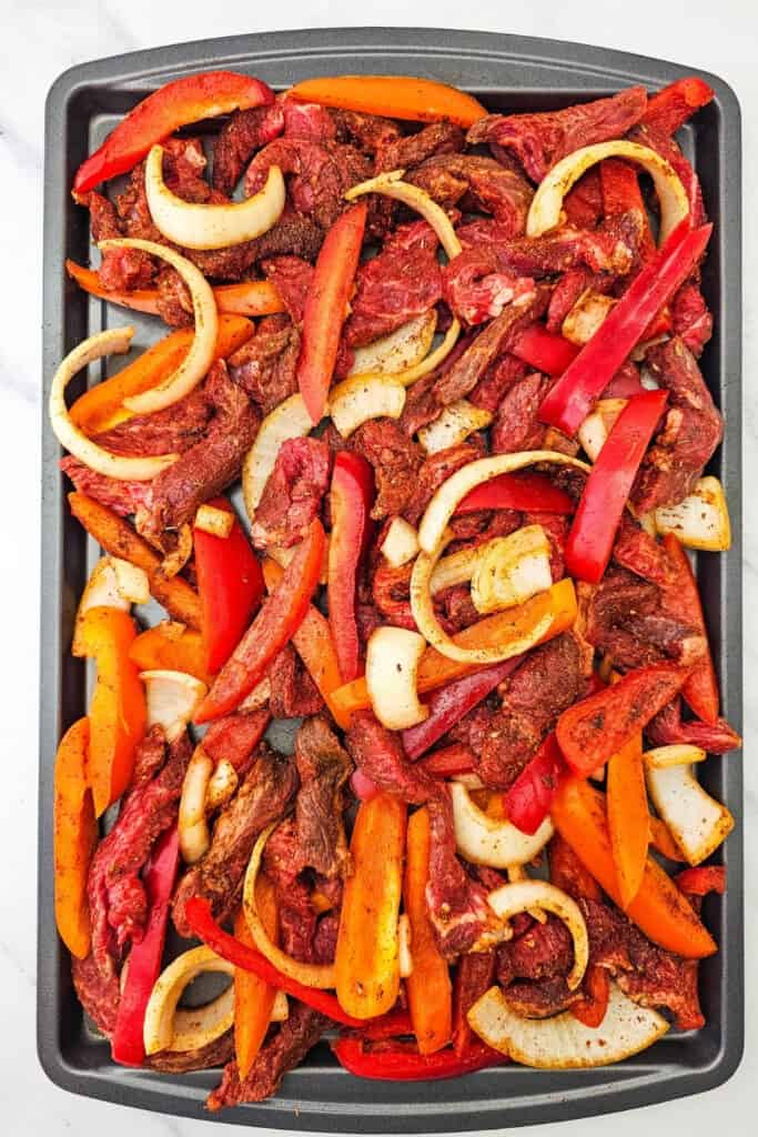 Seasoned steak strips are spread across a sheet pan with sliced red and orange bell peppers and white onions. The uncooked steak fajitas are arranged in a single layer and ready to bake for a quick 30 minute meal.