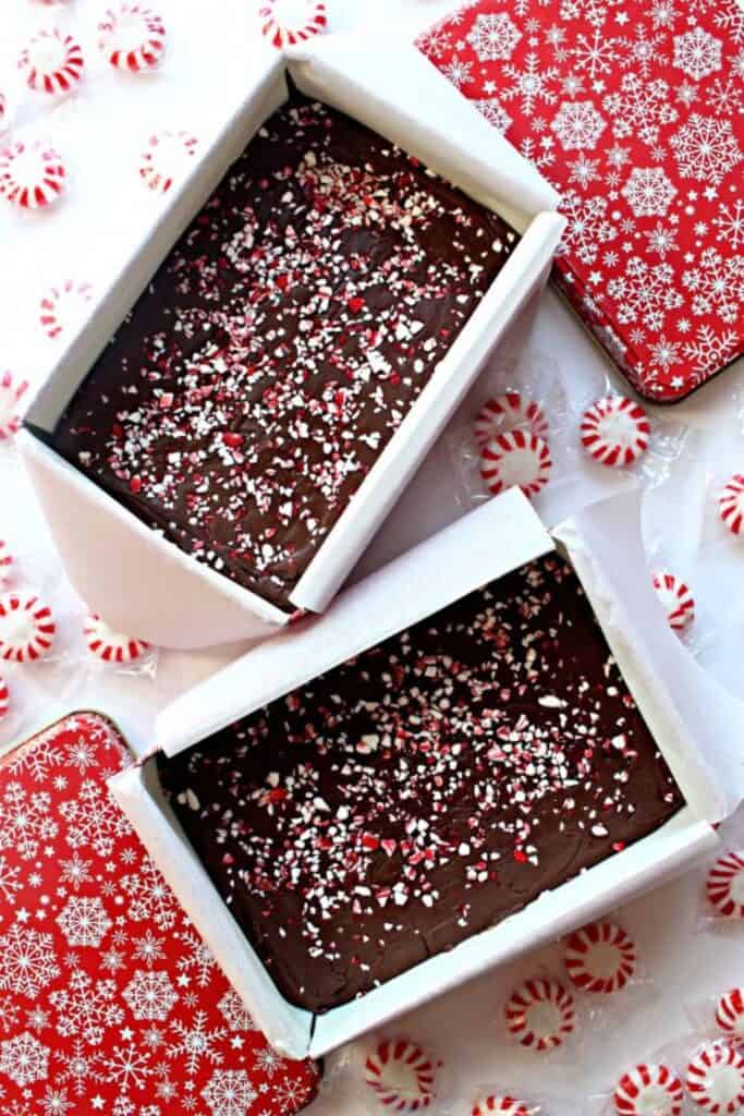 Two gift tins filled with rich chocolate fudge topped with crushed peppermint candy, surrounded by red and white striped peppermint discs. The tins have festive red lids with white snowflakes, making this a classic Christmas dessert tray treat.