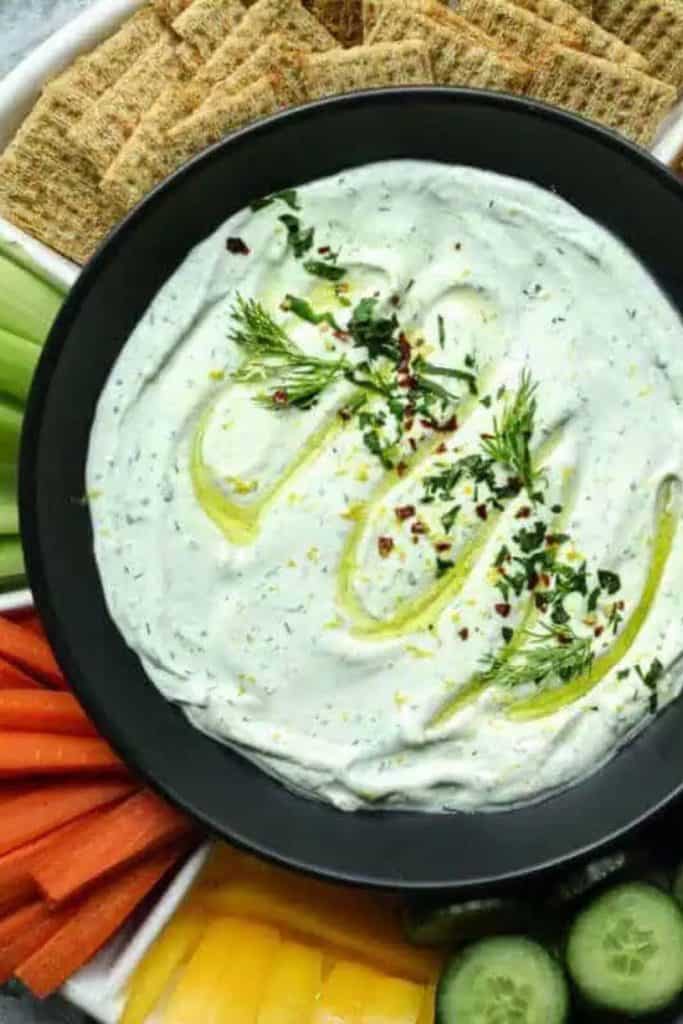 Creamy Ranch Cottage Cheese Dip served in a black bowl, topped with fresh dill, herbs, and a drizzle of olive oil. Surrounded by crackers, carrots, celery, cucumbers, and bell peppers.