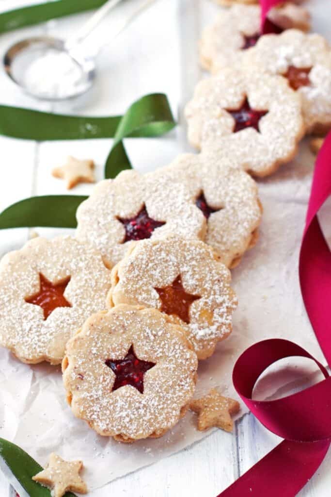 Linzer cookies dusted with powdered sugar are filled with vibrant red raspberry and orange jam, peeking through star-shaped cutouts in the top cookie layer. Arranged with festive ribbons and mini star cookies.
