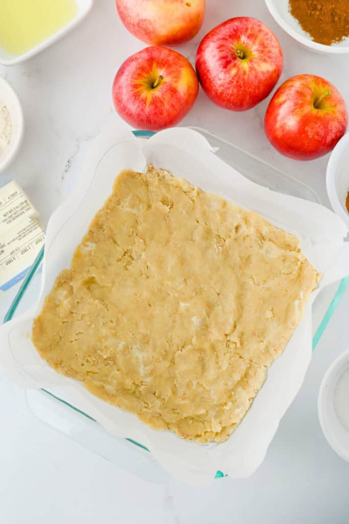 Apple pie bar crust pressed evenly into a parchment lined square baking dish before adding the apple filling. Fresh red apples and baking ingredients surround the pan during the preparation process.