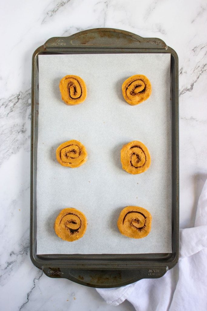 Six unbaked pumpkin cinnamon roll cookies arranged on a parchment-lined baking sheet, spaced evenly and ready for the oven. The swirled cookie dough showcases visible cinnamon sugar spirals against the orange dough.