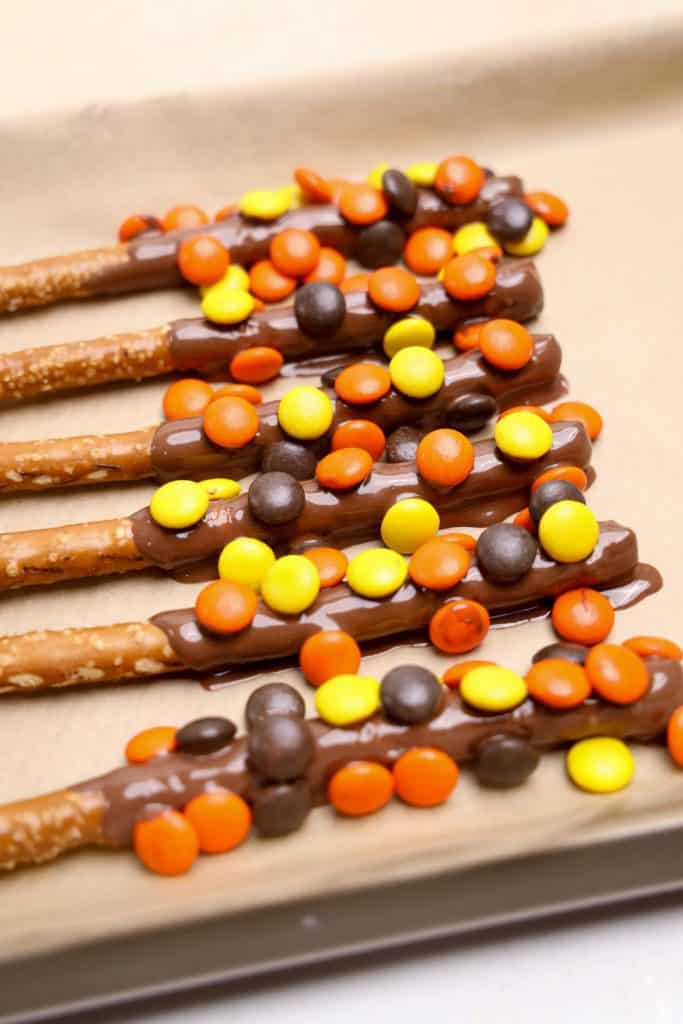 Reese's fall dipped pretzels lined up on parchment paper, coated in glossy melted chocolate and topped with orange, yellow, and brown Reese’s Pieces. The festive colors and candy topping give the pretzels a fall-themed twist.