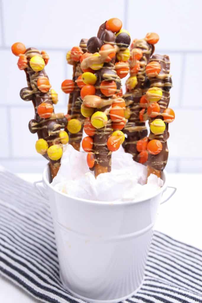 Chocolate-dipped pretzel rods decorated with orange, yellow, and brown Reese’s Pieces and drizzled with peanut butter, displayed upright in a white pail lined with tissue paper.