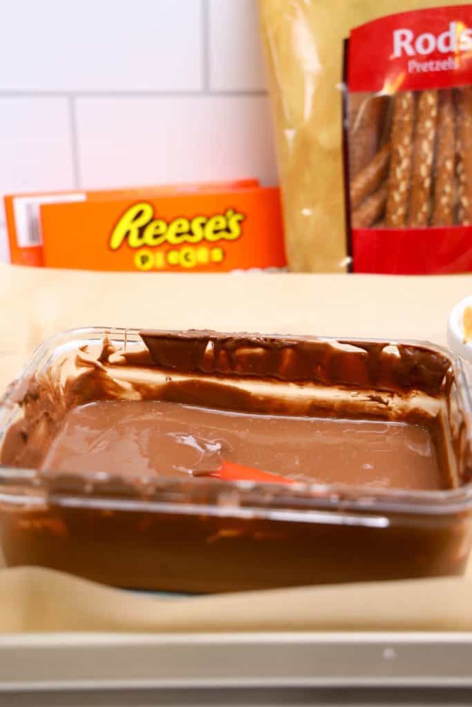 Close-up of Reese's Pretzel Rods ingredients, including a container of melted chocolate with a spatula, a bag of pretzel rods, and a box of Reese’s Pieces in the background. These are the key items for assembling the sweet and salty fall treat.