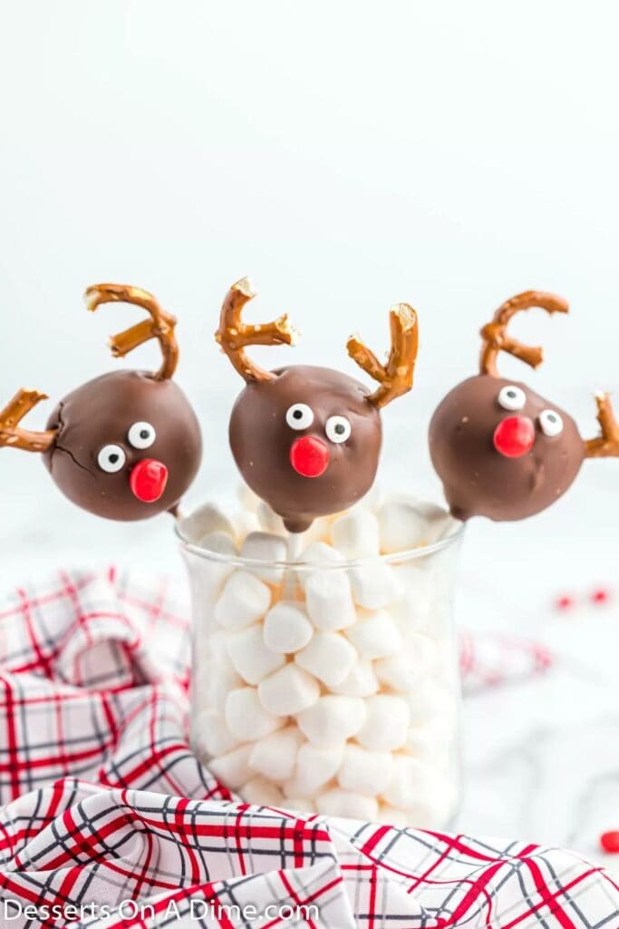 Chocolate cake pops decorated as reindeer feature red M&M noses, candy eyes, and pretzel antlers. They are displayed standing upright in a clear glass filled with mini marshmallows.