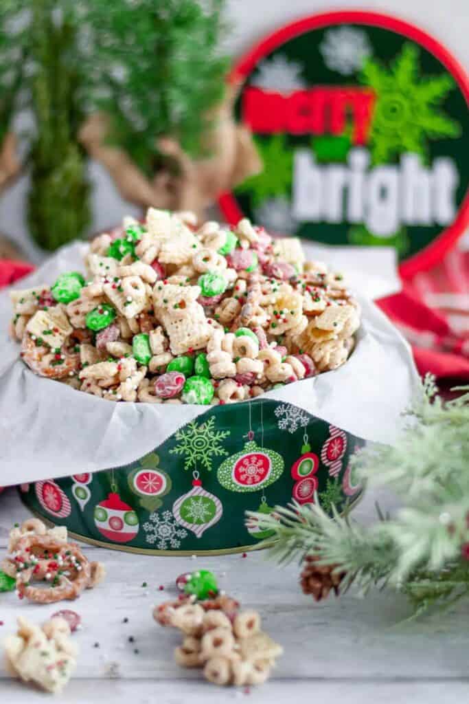 A festive tin filled with Reindeer Chow snack mix made of Chex cereal, pretzels, Cheerios, red and green M&Ms, and holiday sprinkles coated in white chocolate.