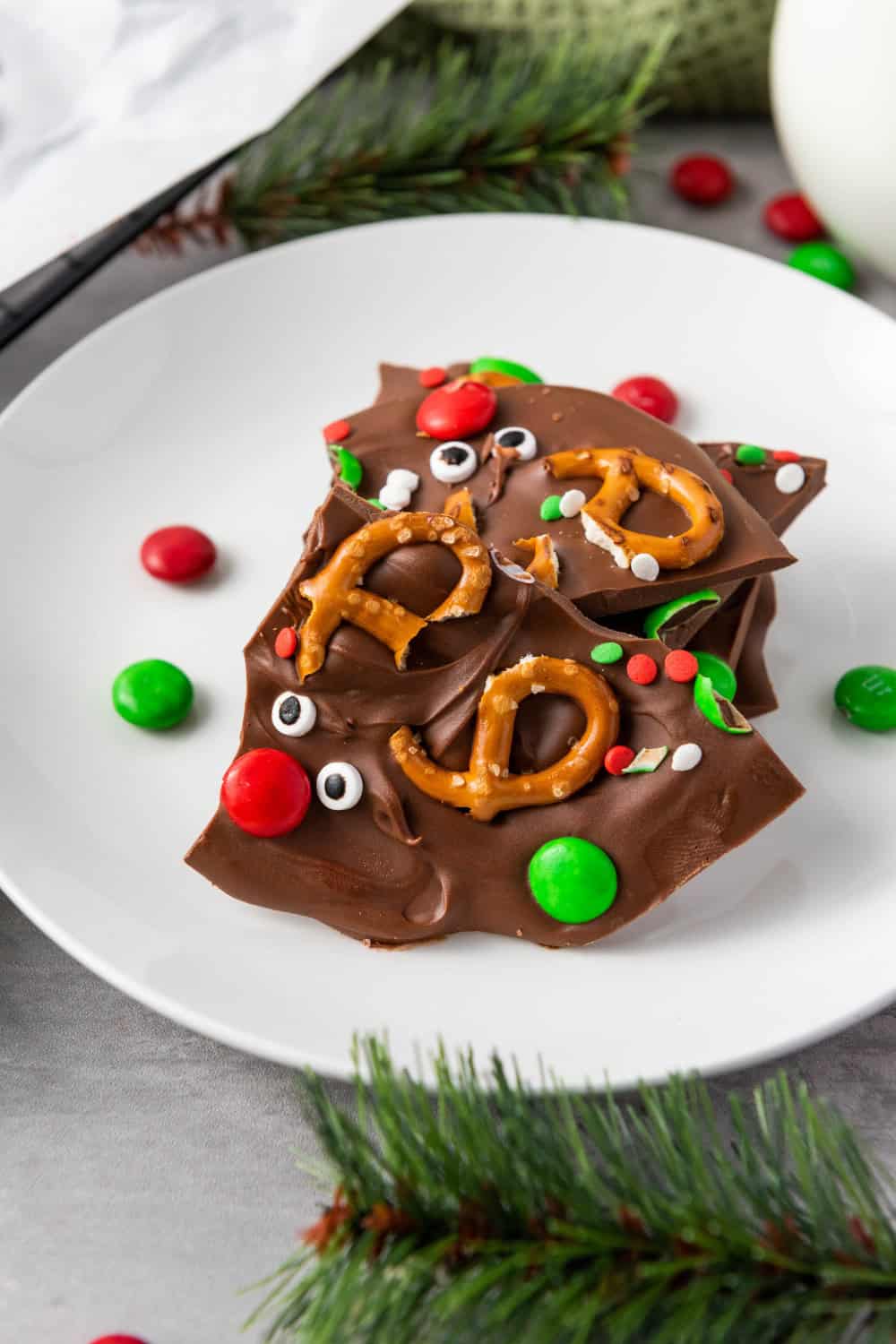 Pieces of Rudolph Christmas Bark decorated with pretzel antlers, candy eyes, red and green candies, and festive sprinkles, served on a white plate.