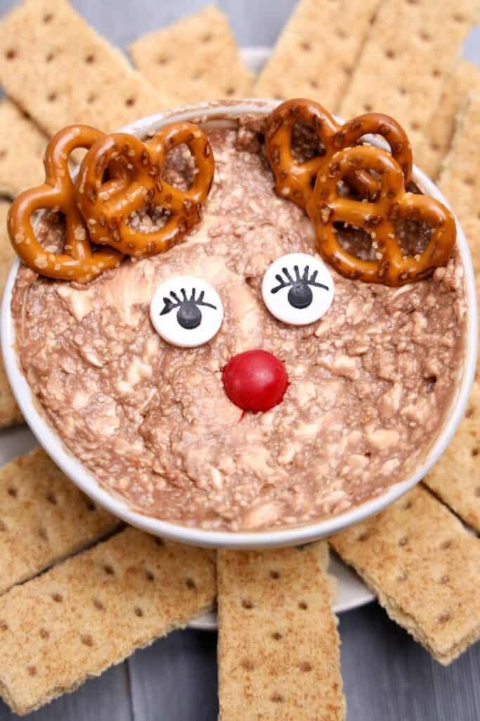Rudolph the Reindeer Christmas Dip in a white bowl, decorated with candy eyes, a red M&M nose, and mini pretzels as antlers. The Nutella and cream cheese mixture is surrounded by neatly arranged graham cracker pieces for dipping.